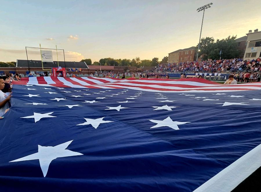 Photo is looking across an oversized American flag being held open on a football field. People are standing around the edges of the flag holding it as spectators in the bleachers watch.