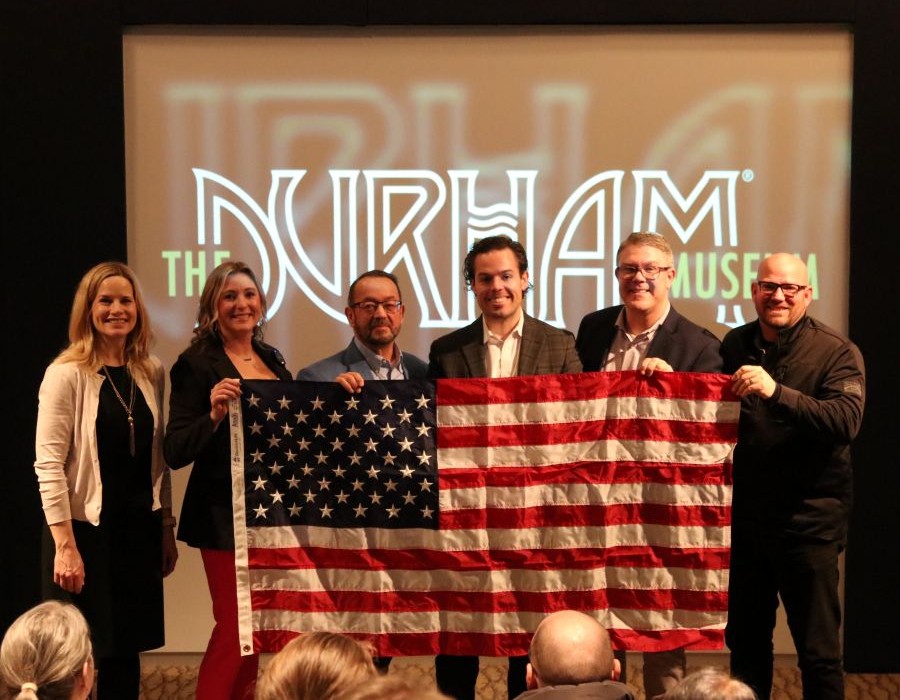 Six people stand together holding a 3-foot-by-5-foot American flag. Written on a projector behind them is "The Durham Museum."