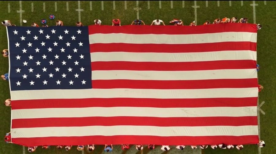 Photo is a drone image looking down on an oversized American flag being held open on display on a football field. People stand around the edges of the flag holding it.