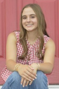 Photo is a senior portrait of Emma B, a WoodmenLife member in Eddyville, KY. She is wearing a red-and-white checkered shirt and jeans, and she is seated in front of a red background.