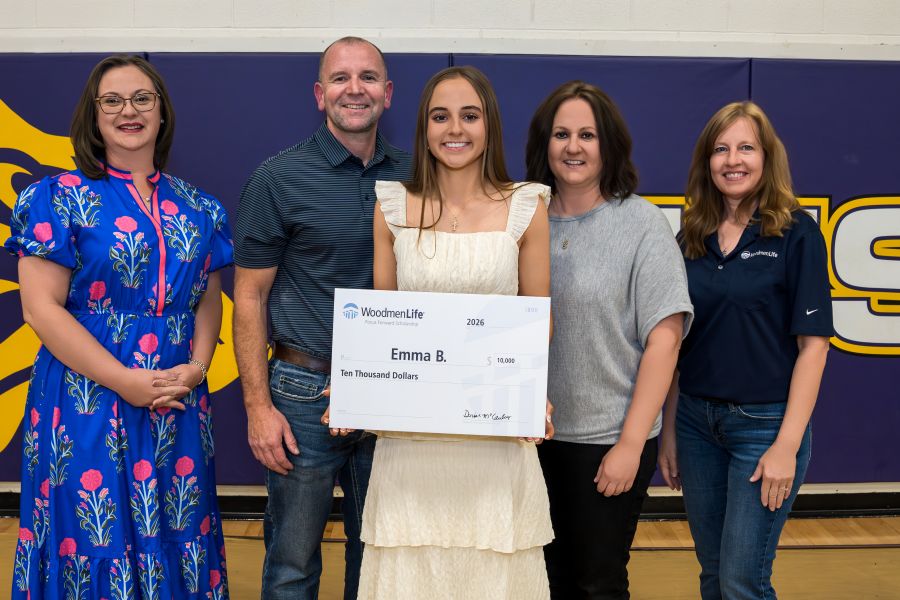 Photo shows five people standing together, posing for the photo. At the center is a young woman holding an oversized WoodmenLife-branded check. The check is made out to Emma B. in the amount of $10,000. Flanking the young woman are a man and woman, her parents. The last two people -- two women on either end of the photo -- are WoodmenLife associates who presented the scholarship check.