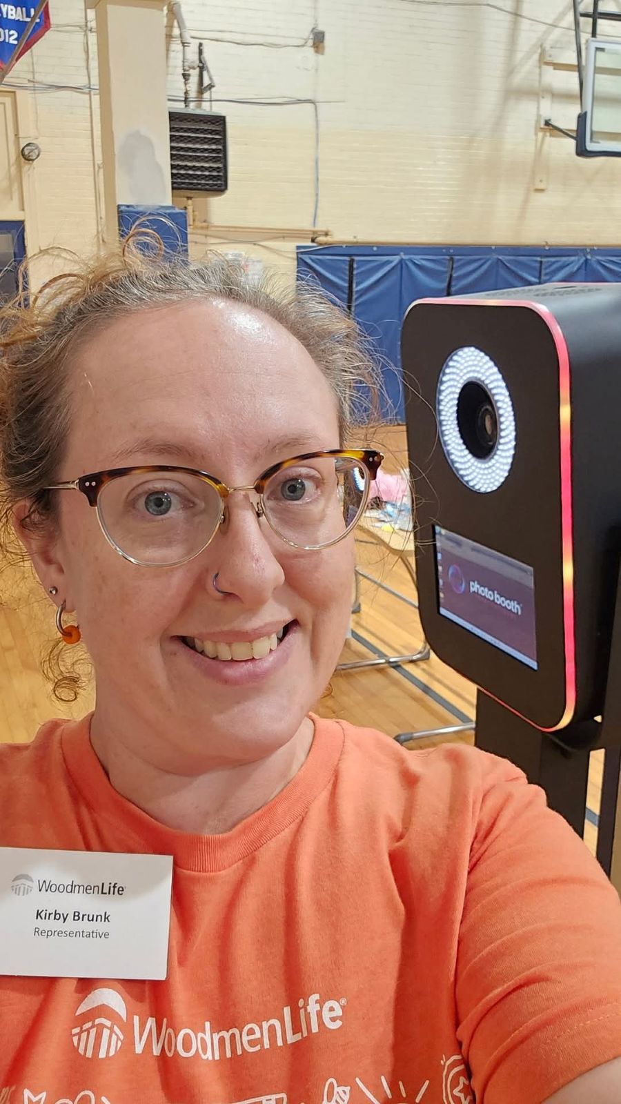 Photo is a selfie of a woman. She is wearing an orange T-shirt and a name tag that says "WoodmenLife, Kirby Brunk, Representative."