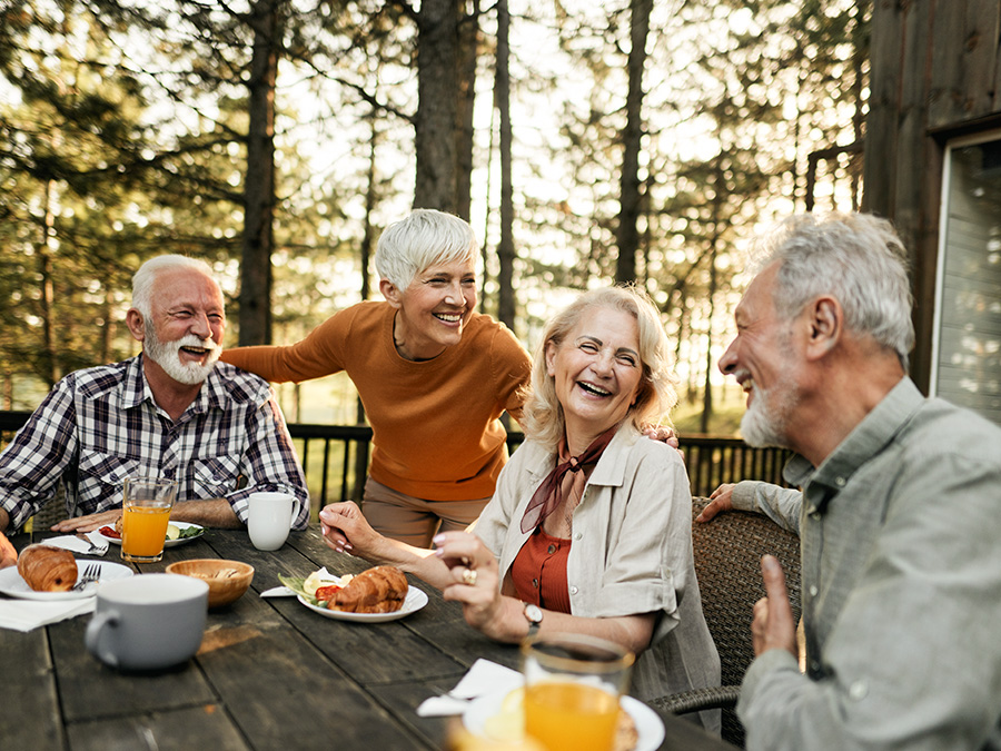 Stock photo shows a group of four older adults sitting at a table outside. There is food on the table in front of them and tall trees behind them. They are all smiling at one another.