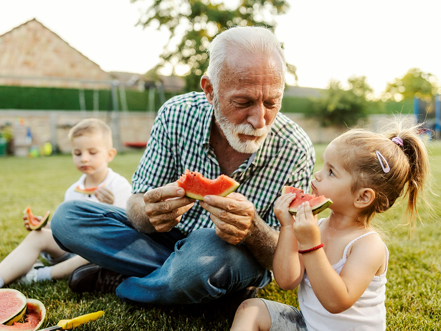 Stock photo shows an older man sitting on the grass with a young girl on one side of him and a young boy on the other. All three are eating slices of watermelon.