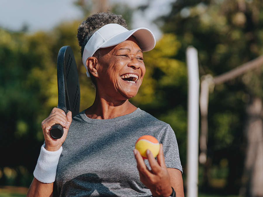 Stock photo shows an older woman holding a tennis racket and tennis ball. She is laughing as she looks into the distance.