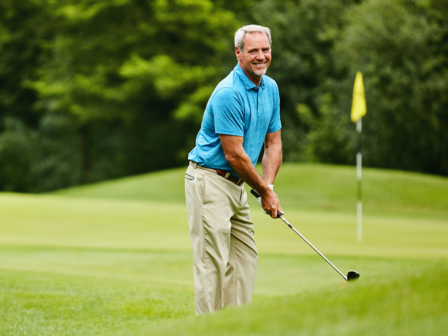 Stock photo shows an older man on a golf course. He is holding a club, and there is a golf flag behind him. He is smiling at the camera.