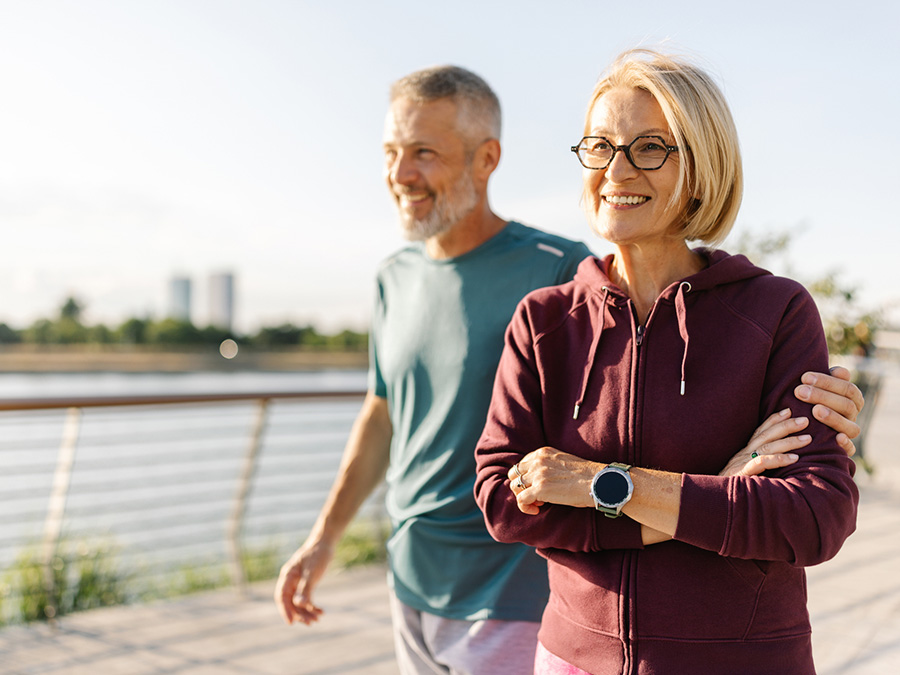 Stock photo shows an older man and woman walking. The man has his arm around the woman. Behind them, there is a railing along the walkway that they're on; beyond the railing is a body of water.