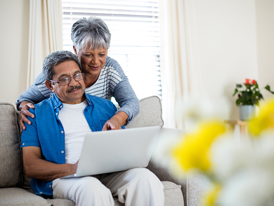 Stock photo shows a man sitting on a couch with a laptop open on his lap. A woman is standing behind him, leaning over, and pointing at something on the laptop.