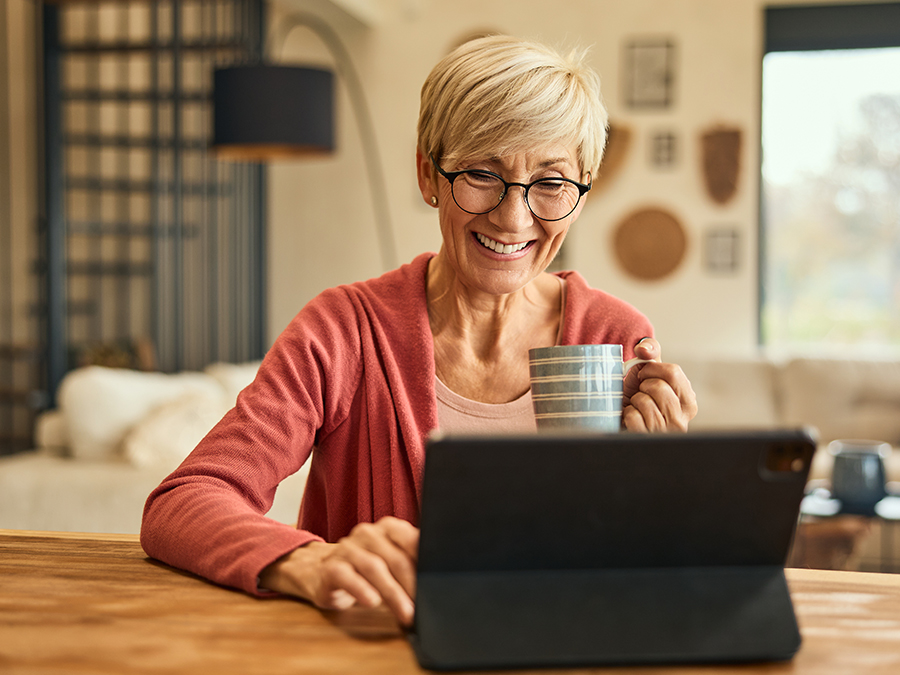 Stock photo shows an older woman sitting at a table, holding a coffee cup and looking at a tablet. She is smiling.