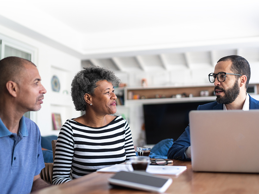 Stock photo shows a man and a woman sitting at a table with another man. There is an open laptop and paperwork on the table.
