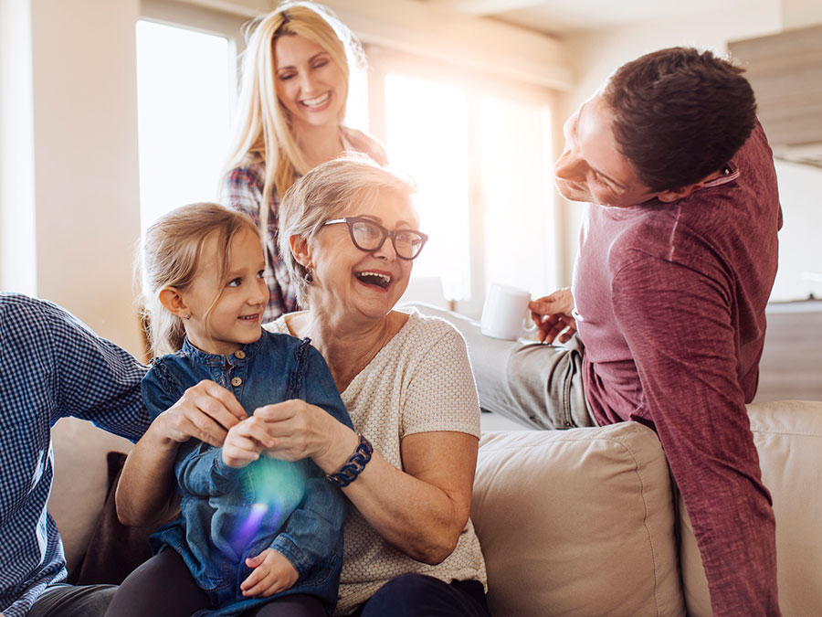 Stock photo shows an older woman sitting on a couch holding a young girl on her lap. Behind her, sitting on the back of the couch and looking over, are two young adults. They are all happy and smiling with one another.