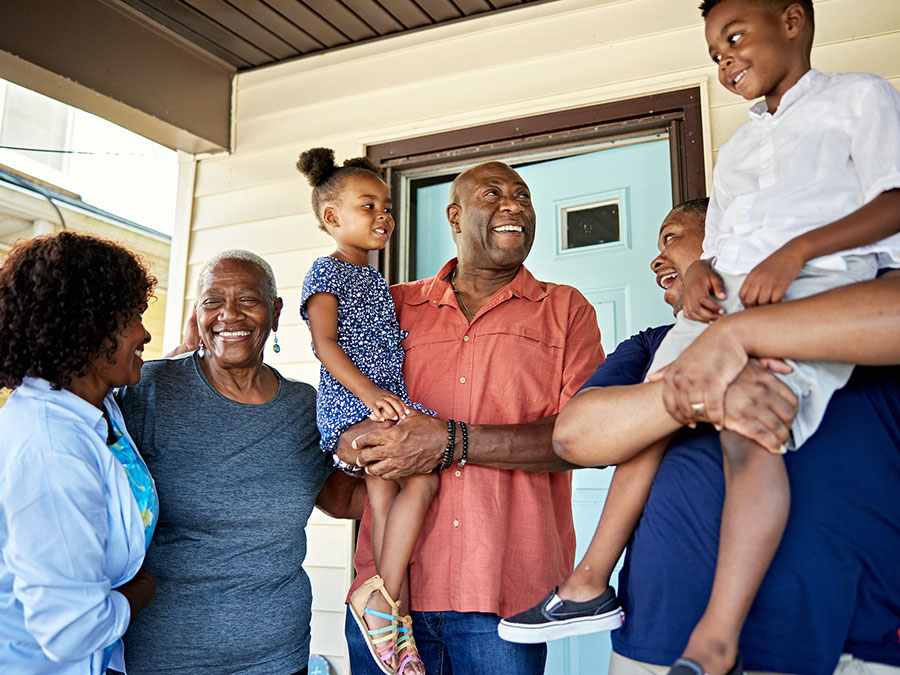 Stock photo shows a family standing on a porch. There are four adults -- two older adults and two younger adults. The older man and younger man are both holding a child in their arms. Everyone is happy and smiling.