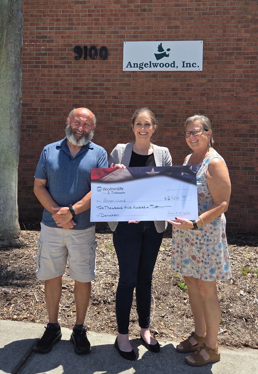 Photo shows three people standing together and holding an oversized check. The check is made out to Angelwood in the amount of $2,500. Behind them is a brick wall with the Angelwood, Inc. logo.
