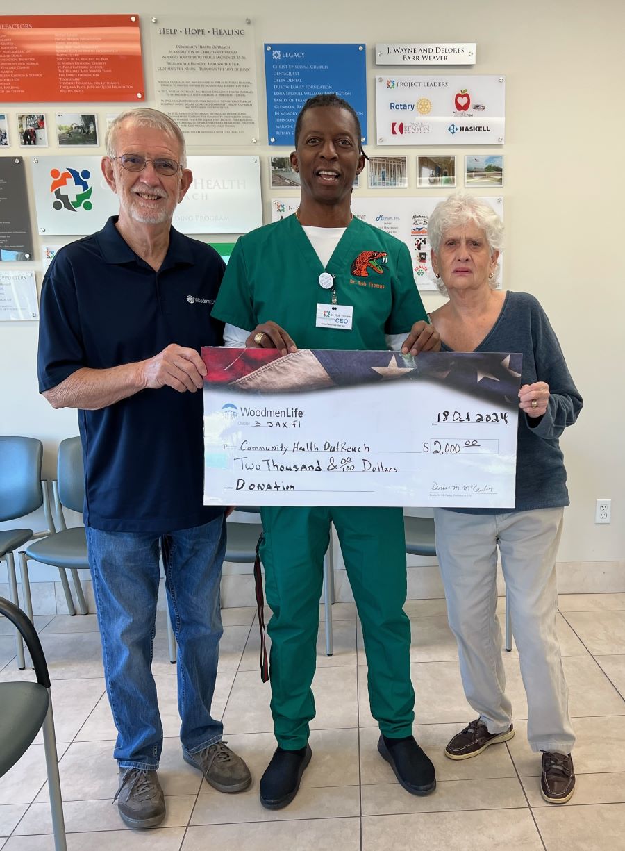 Three people stand holding an oversized check made out to Community Health Outreach in the amount of $2,000. The middle person is wearing medical scrubs.