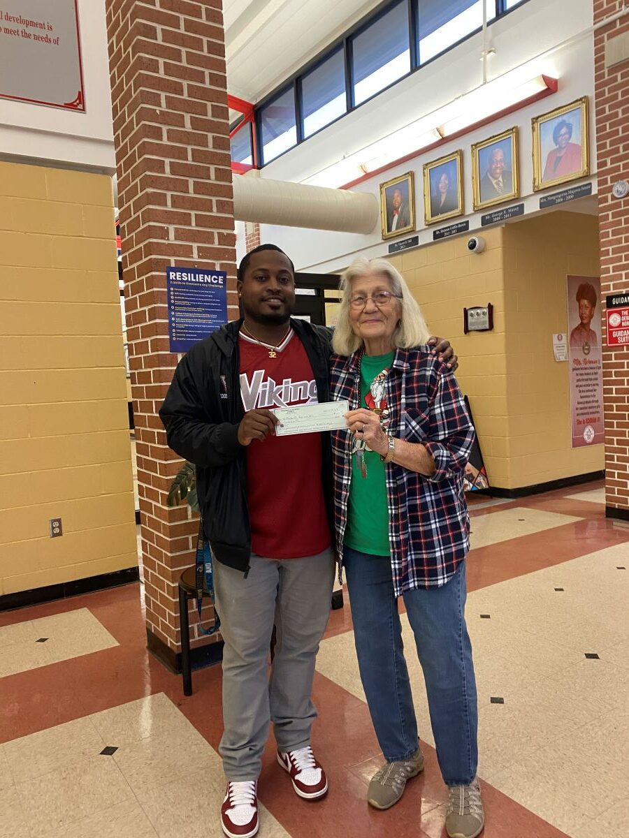 Two people — a man and a woman — stand together holding a check. They are standing in a school hallway.