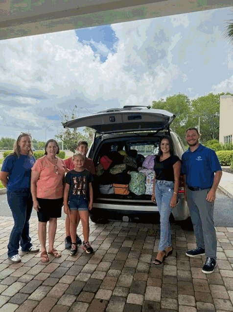 Six people stand around the open hatchback of a vehicle. In the back of the vehicle are stacks of backpacks filled with donated school supplies.