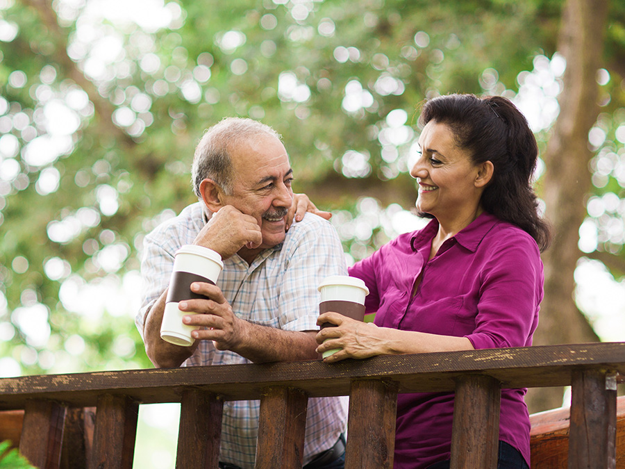 Stock photo shows a man and a woman. They are standing outside, leaning over a wooden railing, and holding coffee cups. They are smiling at each other lovingly.