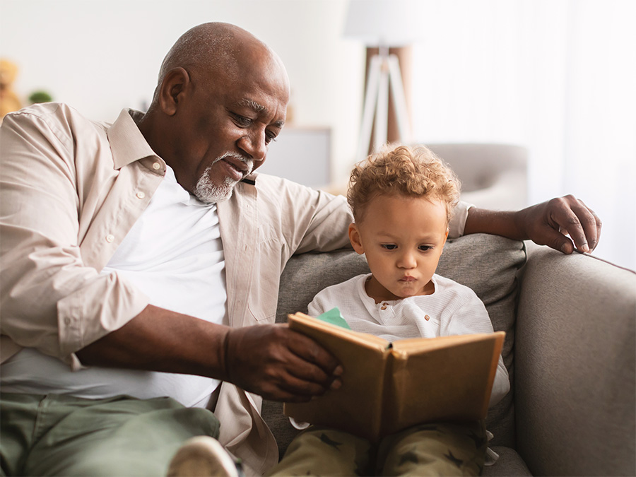 An older man and a young boy sit together on a couch. They are both looking at a book the man is holding in front of the boy.