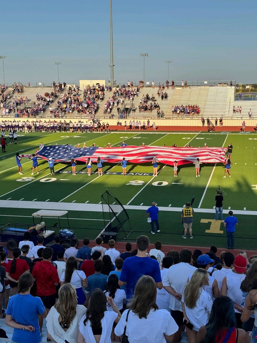Photo shows an oversized American flag being displayed on a football field. People are standing around the flag holding it open as spectators watch from the near bleachers and from the opposite side of the field.