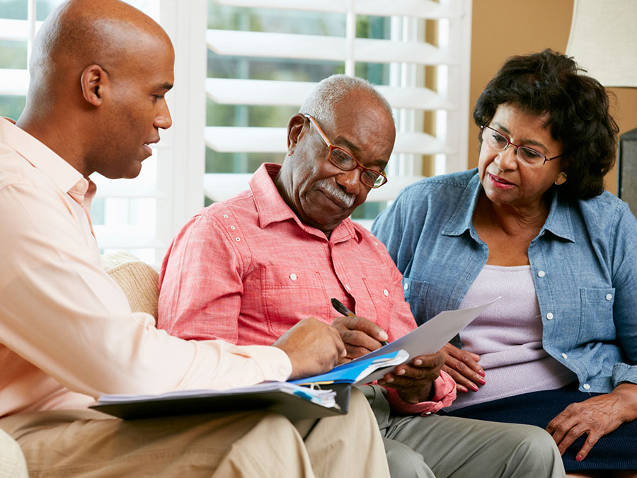 Stock photo shows an older couple sitting with a professional. The professional is showing them paperwork, and the man is signing a piece of paper.