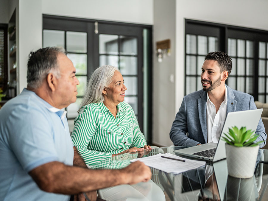 Stock photo shows an older couple sitting at a table with a professional. There is a laptop open in front of them and papers on the table.