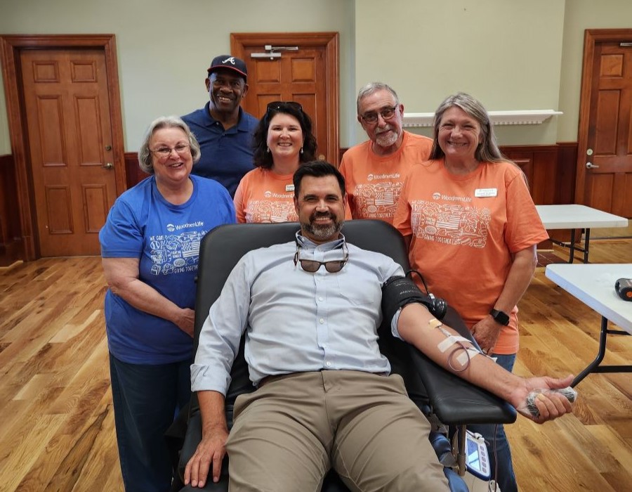 A man is propped up on a table as he donates blood. Five WoodmenLife members stand around him.