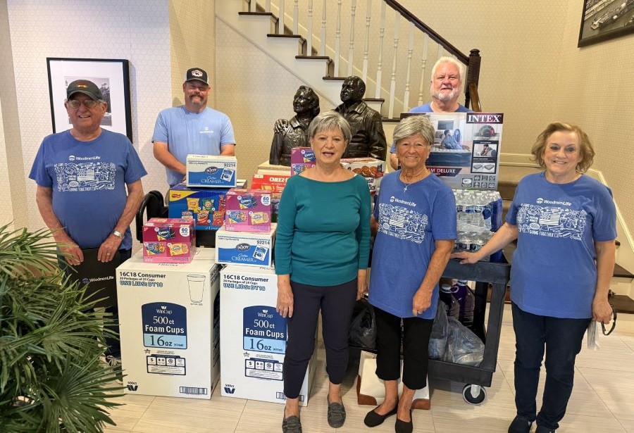 Six people stand around stacks of donations. Among the donations are boxes of foam cups, boxes of chips and cookies, and packs of bottled water. Behind the group is a statue of two people, and behind the statue is a set of stairs going up.