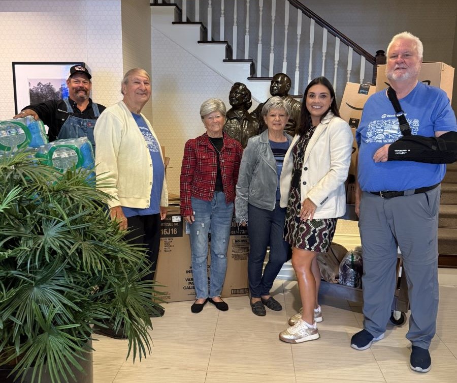 Six people stand together in an entryway. There is a plant to the left of them. Behind them are stairs going up and a statue of two people.