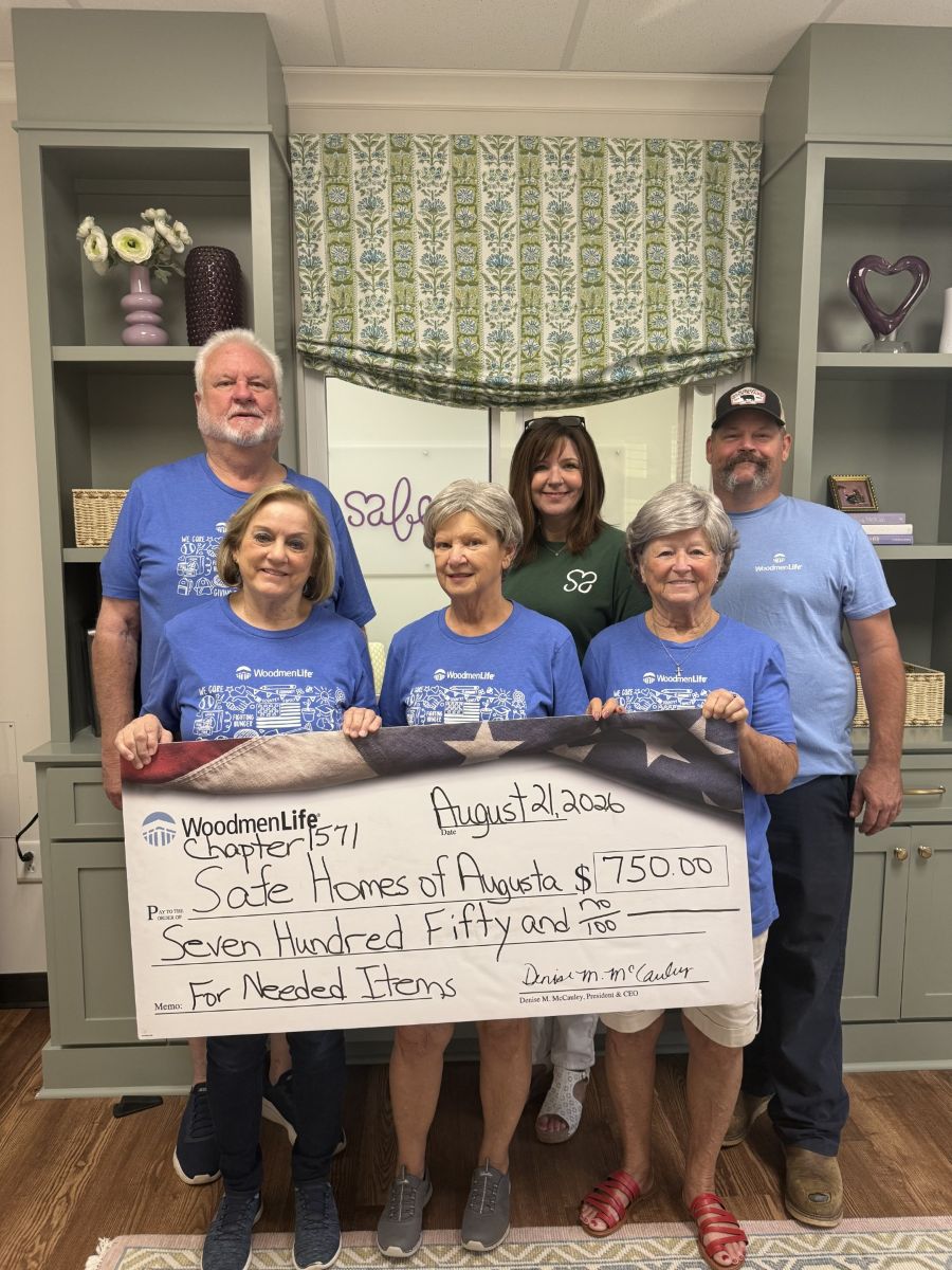 Photo shows six people standing together with an oversized check. The check is made out to SafeHomes of Augusta in the amount of $750. The group is standing indoors, and there are shelves and a window behind them.