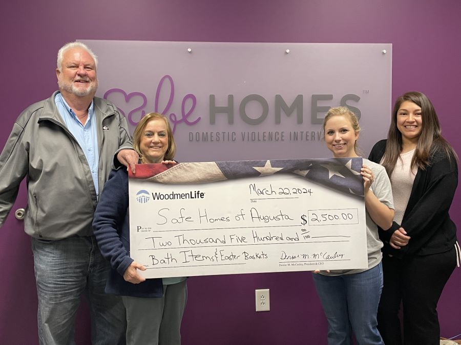 Four people hold an oversized check. The check is for $2,500 and is made out to Safe Homes of Augusta. The group is standing in front of a purple wall with the "Safe Homes" logo.