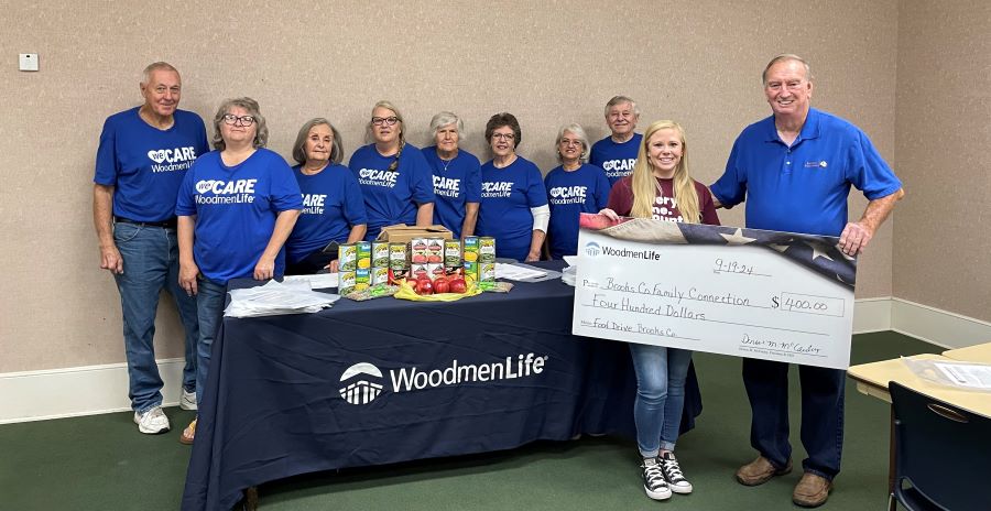 Ten people appear in this photo. The two people in the foreground are holding an oversized check for $400 made out to Brooks Co. Family Connection. The eight people in the background are standing behind a WoodmenLife-branded table and are all wearing blue T-shirts that say "WeCare WoodmenLife."