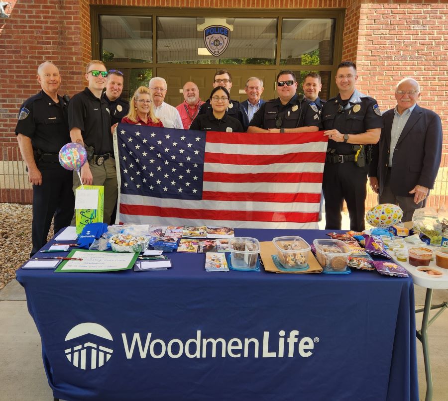 Thirteen people stand together holding a U.S. flag. Several of the people are police officers in uniform. In front of the group is a table with a WoodmenLife-branded table cloth. On top of the table are WoodmenLife brochures, koozies and a variety of food.