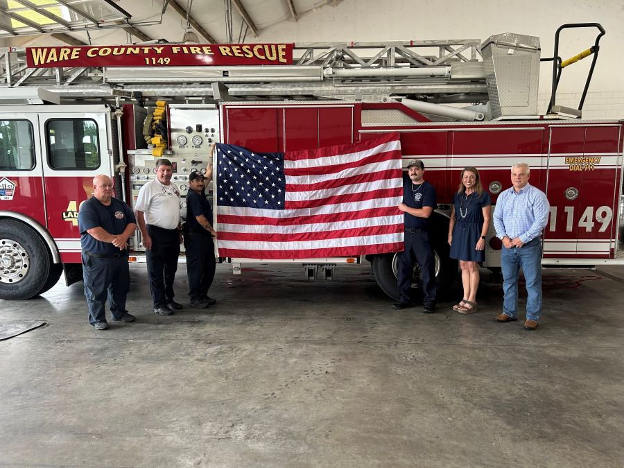 Six people stand together in the photo, with the center two people holding up an American flag. The group is standing alongside a fire truck.