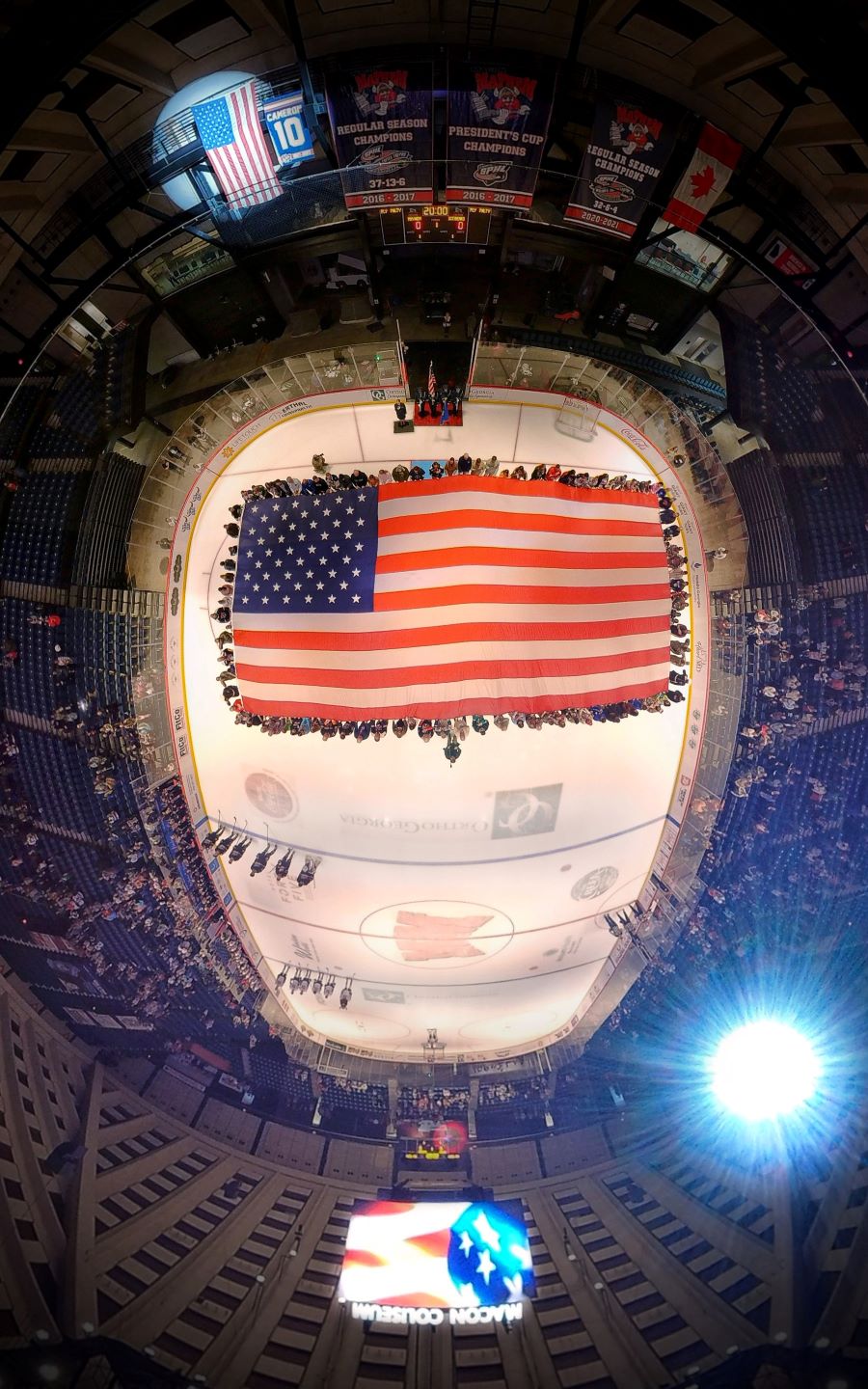 Photo is a drone image from high above an ice hockey rink. On the ice is an oversized American flag, and volunteers are standing around the edge holding the flag. There's a fisheye lens effect on the photo.