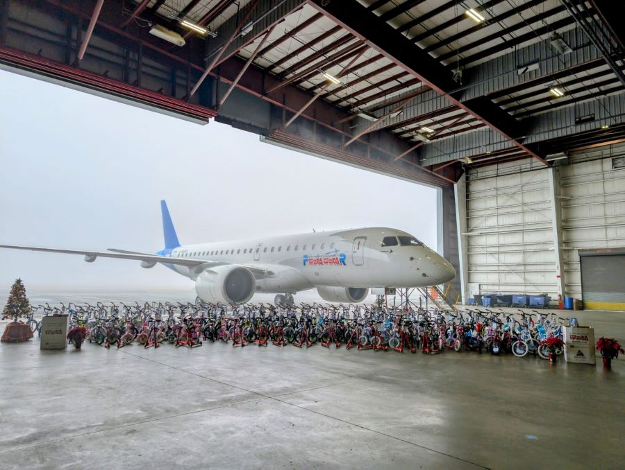 An airplane sits in a hangar. Lined up in front of the plane are dozens of children's bicycles. On the ends of the rows of bicycles are a small Christmas tree, a poinsettia, and boxes with the Toys for Tots logo.