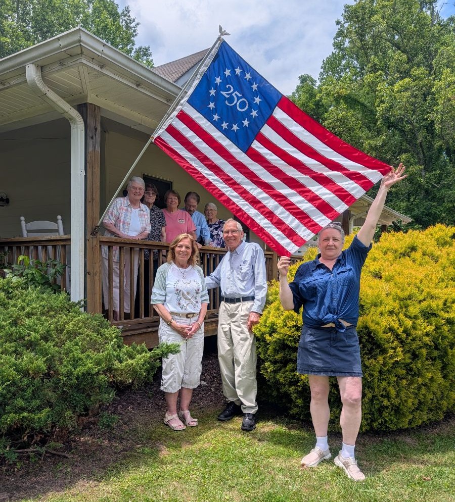 A group of eight people stand together in the photo. Five of the people are standing on a deck, and the other three are standing below on the ground. A commemorative America 250 flag is affixed to the deck, and one of the people is holding the flag out. There are green bushes, trees, and grass around them.