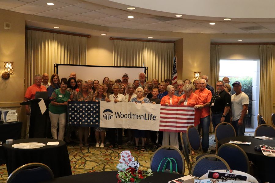 A group of about 40 people stand together for a photo. The front row of people are holding a WoodmenLife banner that is decorated with stars and stripes on either end.