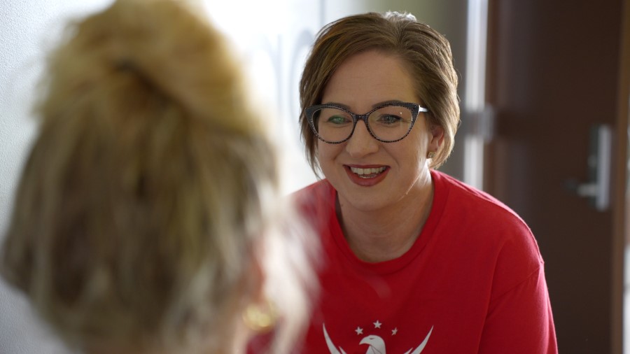 Photo shows a woman smiling. She is wearing a red T-shirt and looking at someone whose head we can see the back of.