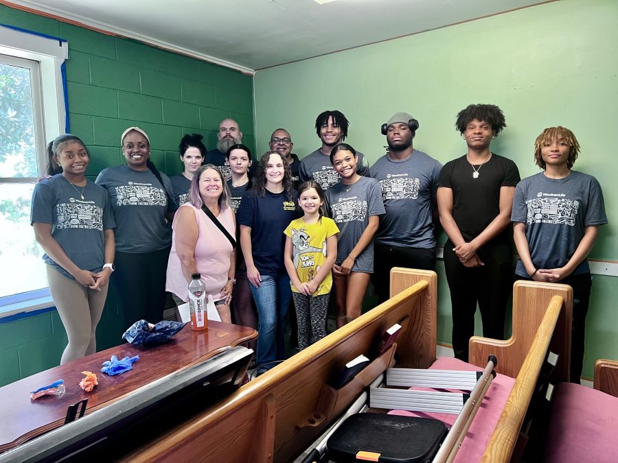 A group of 14 volunteers stand together with their newly painted green wall behind them. Church pews are in the foreground of the photo.