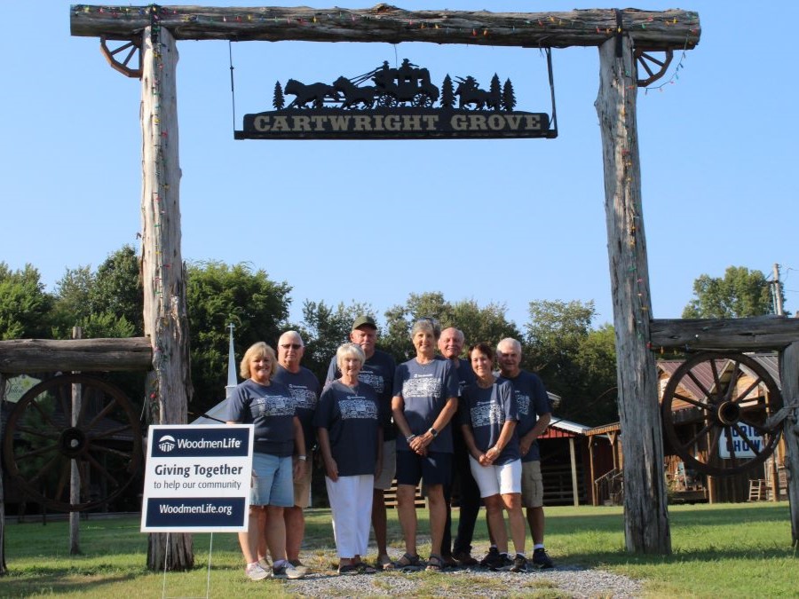 Eight people stand together under a sign that says "Cartwright Grove." They are all wearing blue-gray WoodmenLife-branded t-shirts. Beside them is a sign that says "WoodmenLife. Giving Together to help our community. WoodmenLife.org."