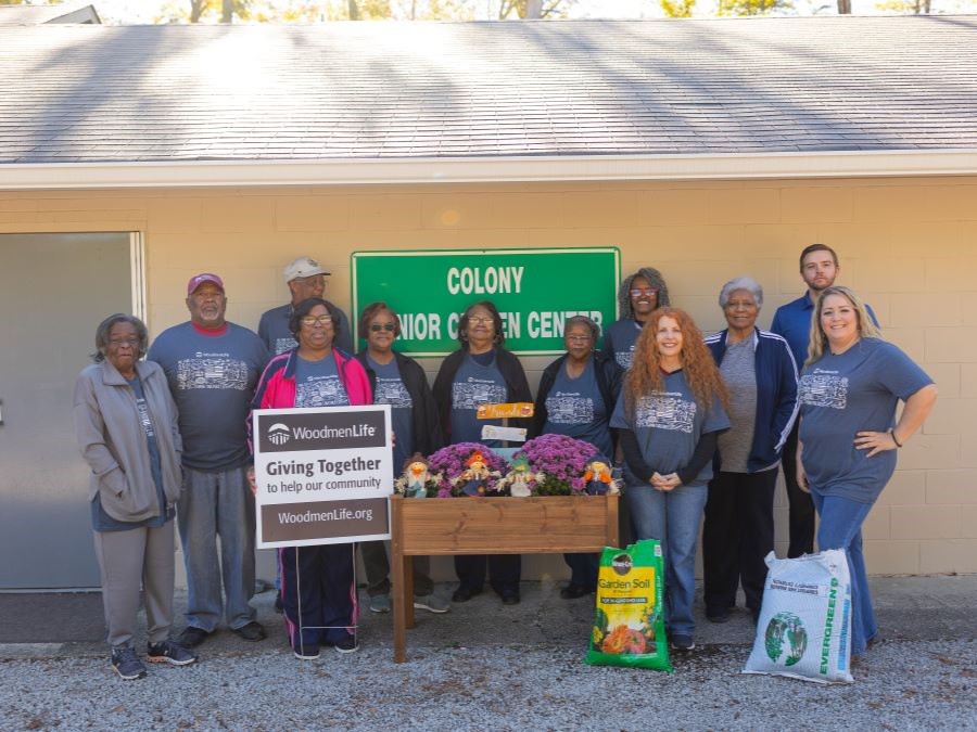 A group of 12 people stand around a newly planted flower bed. One person is holding a sign that says "WoodmenLife. Giving Together to help our community. WoodmenLife.org" Everyone is wearing blueish-gray WoodmenLife-branded t-shirts. On the wall behind them is a sign that says "Colony Senior Citizen Center."