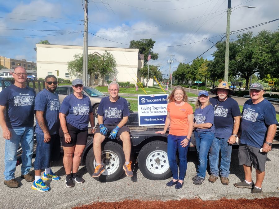 A group of eight people stand together for the picture. In the middle is a sign that says "WoodmenLife. Giving Together to help our community. WoodmenLife.org." Most of the people are wearing blue-gray WoodmenLife-branded t-shirts. It appears sunny outside, and many people are wearing hats.