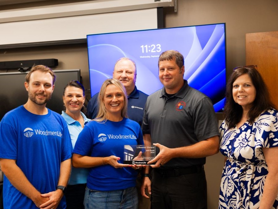 Six people appear in the photo. Two people in the middle are holding a WoodmenLife Heart & Heritage Award, which is small and glass. Two people in the photo are wearing blue WoodmenLife T-shirts.