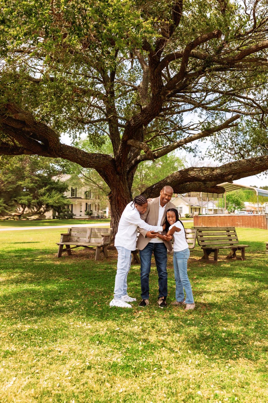 A man and his two children -- a son and a daughter -- stand together hugging. They are below a large tree and are surrounded by green grass.