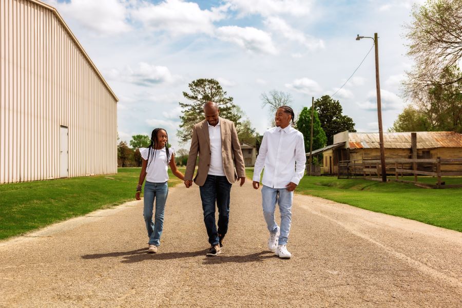 A man walks down a gravel road with his son on one side and his daughter on the other. There is a cloudy blue sky above them.