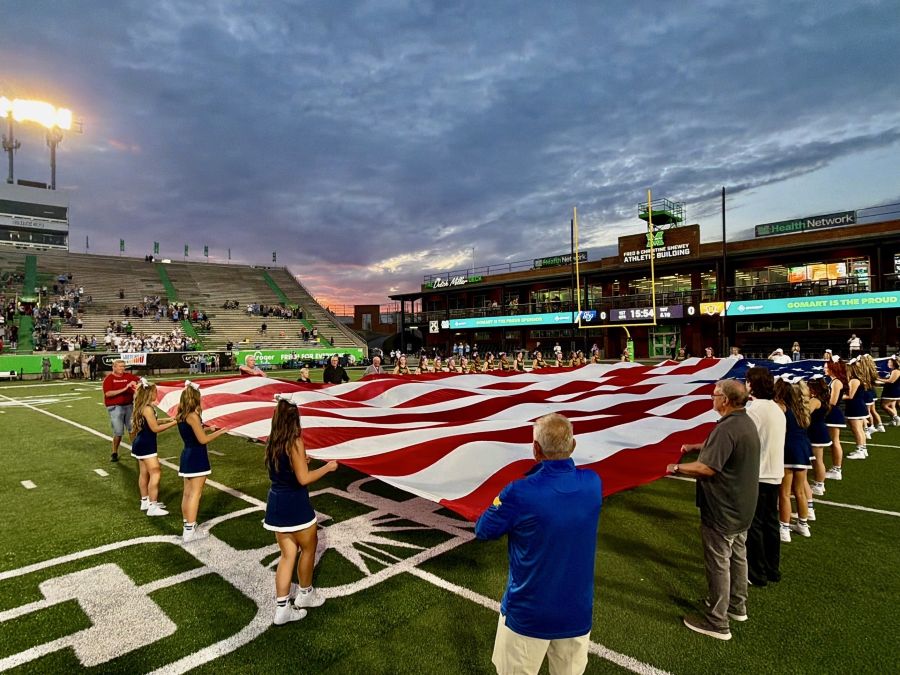Photo shows a mixture of adults and teenage cheerleaders holding open an oversized American flag. They are standing on a football field, and the sun is setting behind them.