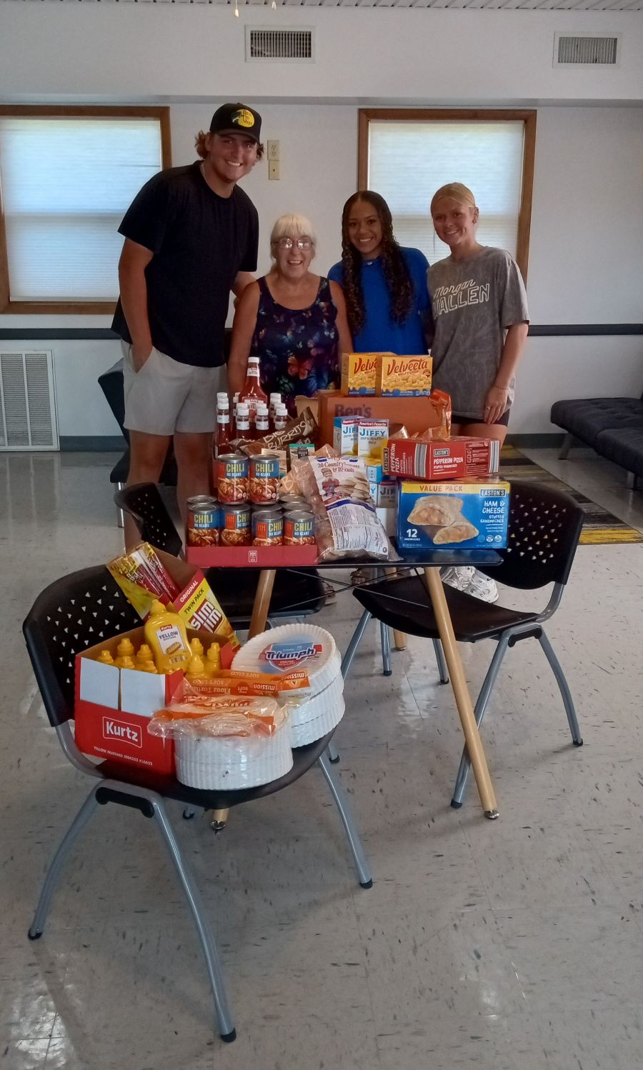Photo shows four people standing behind a table and chairs stacked high with donated food.