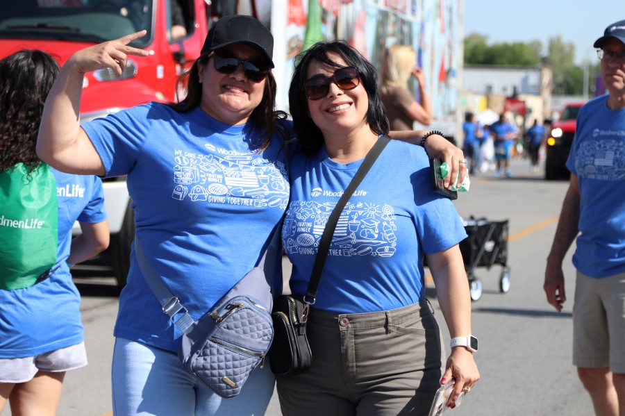 Photo shows two women standing together for the photo. They are both wearing blue WoodmenLife T-shirts. They are both wearing sunglasses in the bright sunlight, and the woman on the left is also wearing a hat. They have an arm around each other, and the woman on the left is holding up two fingers like a peace sign.