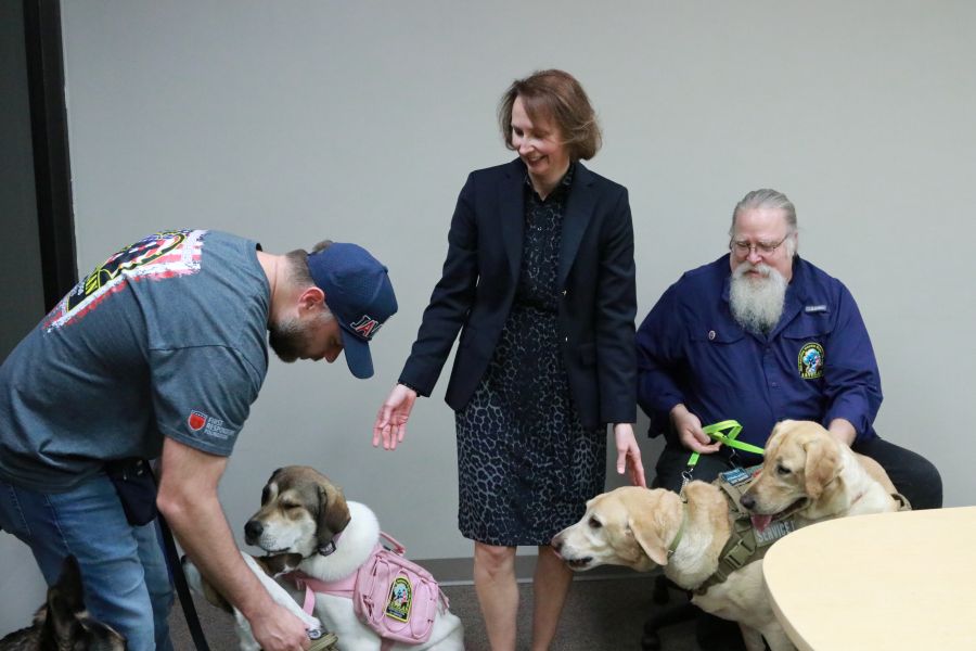 A woman stands in the middle of the photo. On either side of her is a man holding dogs on leashes. The woman is leaning down to pet the dogs.