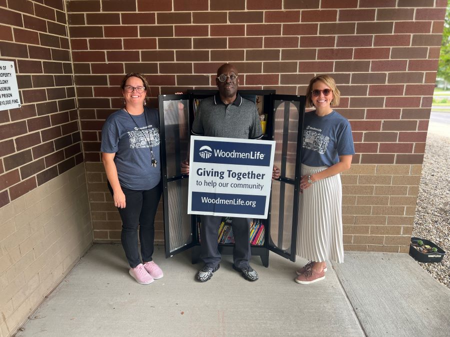 Three people stand in front of the new little free library, which has its door swung open. The middle person is holding a sign that says "WoodmenLife. Giving Together to help our community. WoodmenLife.org."